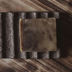 Puede incluir: Una pastilla de jabón marrón descansa sobre un jabonera gris oscuro con ranuras. El jabón tiene forma cuadrada y es de color marrón claro con vetas más oscuras. La jabonera es rectangular y está hecha de un material texturizado.