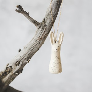A ceramic hare decoration with hand-drawn black eyes and a black nose. The ornament is white with black markings and is hanging from a gold string.