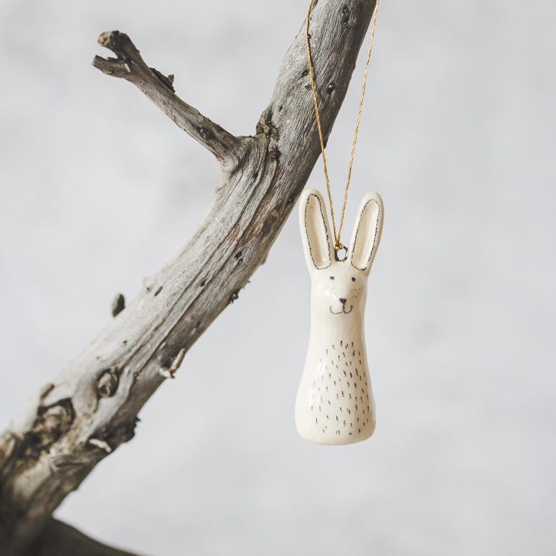 A ceramic hare decoration with hand-drawn black eyes and a black nose. The ornament is white with black markings and is hanging from a gold string.