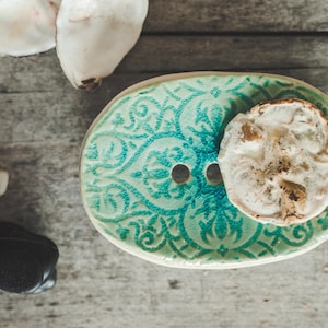 May include: A ceramic soap dish with a teal and white floral design. The dish has a small hole in the center and a white, round soap bar is resting on the dish.