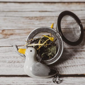 May include: A ceramic duck tea infuser with a stainless steel mesh basket, filled with loose tea leaves and flower petals. The duck has a yellow beak and is attached to a chain. The infuser is open, revealing the tea inside.