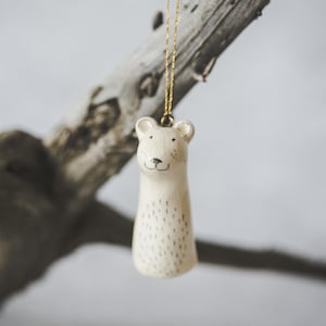 A ceramic bear decoration with hand-drawn black eyes and a black nose. The ornament is white with black markings and is hanging from a gold string.