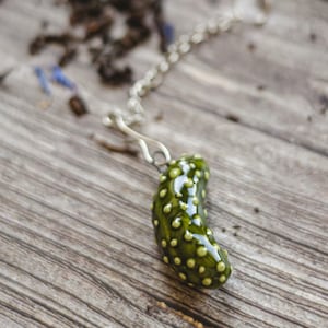 Könnte beinhalten: Ein grüner, keramik-tee-infuser in Form einer Gurke mit weißen Punkten. Der Infuser ist an einer silbernen Kette befestigt.