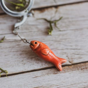 Pode incluir: Infusor de chá de cerâmica com um design de peixe laranja brilhante. O infusor em forma de peixe é preso a um coador de chá de aço inoxidável com uma corrente. O fundo é uma superfície de madeira clara.