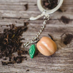 May include: A peach-shaped tea infuser with a green leaf charm and a silver chain. The infuser is on a wooden surface with loose leaf tea scattered around it.