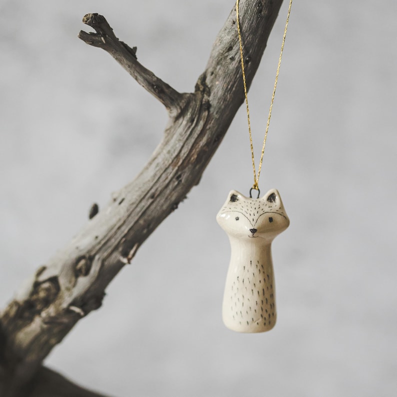 A ceramic racoon decoration with hand-drawn black eyes and a black nose. The ornament is white with black markings and is hanging from a gold string.