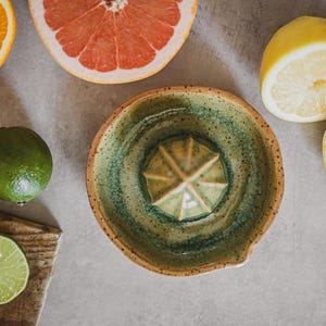 May include: A green and brown ceramic citrus juicer with a star-shaped reamer. The juicer is surrounded by various citrus fruits, including oranges, lemons, and limes.