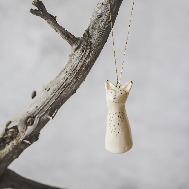 A ceramic fox decoration with hand-drawn black eyes and a black nose. The ornament is white with black markings and is hanging from a gold string.