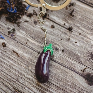 Könnte beinhalten: Ein lila auberginenförmiger Tee-Infuser mit grünem Stiel und Blättern. Der Infuser ist an einer silbernen Kette befestigt und hängt an einem silbernen Teesieb.