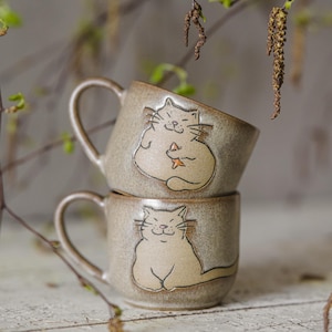 Może przedstawiać: Dwa ceramiczne kubki ułożone jeden na drugim z jasnobrązową glazurą. Każdy kubek ma fantazyjną ilustrację kota w jaśniejszym odcieniu brązu. Koty uśmiechają się i trzymają gwiazdę. Kubki mają zakrzywione uchwyty.