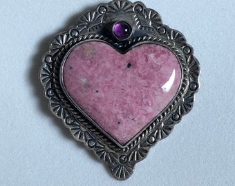 May include: A heart-shaped pendant with a pink stone center and a silver-colored, ornate frame. A small, round, purple stone is set at the top. The pendant has a detailed, textured design around the edges.