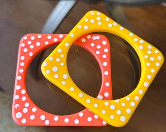 May include: Two square bangles. One is orange with white polka dots, and the other is yellow with white polka dots. The bangles are made of a glossy material and are stacked on top of each other.