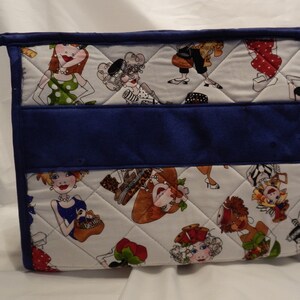 May include: A white quilted zippered pouch with a blue fabric panel and a handle. The pouch features a colorful print of women in various poses and outfits.