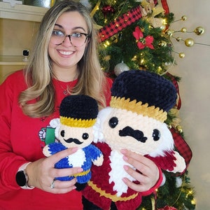 May include: Two crocheted nutcracker dolls. One is red and white with a black hat and the other is blue with a black hat. The dolls are being held by a person.