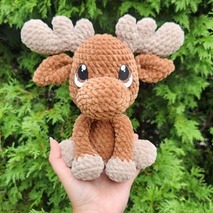 May include: A handmade, plush moose toy in shades of brown and tan. The moose has large, expressive eyes, a soft, textured body, and detailed antlers. The toy is held against a backdrop of green foliage.