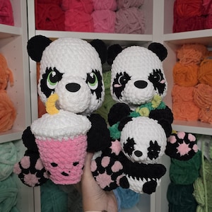 May include: Three crocheted panda bears, one holding a pink and white crocheted cup with a yellow straw, another holding a green and white crocheted flower, and the third sitting on the other panda's lap. The pandas are all black and white with black eyes and noses.