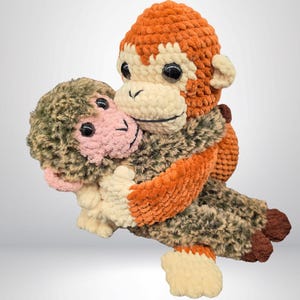 Könnte beinhalten: Zwei handgefertigte Häkelaffen in einer Umarmung. Der größere Affe hat orangefarbenes und cremefarbenes Fell mit schwarzen Augen. Der kleinere Affe hat grünes und rosa Fell. Ein weiches Spielzeug für Kinder.