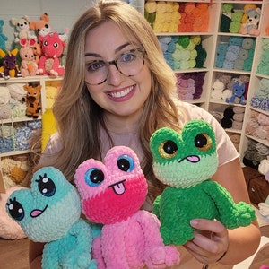 May include: Three crocheted frog plushies in different colors: pink, green, and blue. The frogs have large eyes and are holding their tongues out.