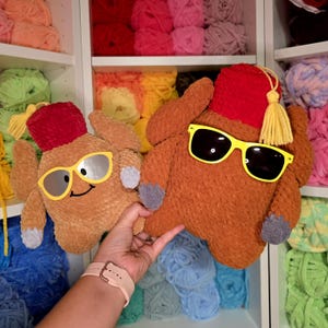 May include: Two plush turkey-shaped pillows. One is light brown with yellow sunglasses and a smiling face. The other is a darker brown with black sunglasses. Both have red fez hats and gray feet.