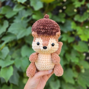 May include: A plush crochet squirrel toy featuring a dark brown textured acorn cap on its head. The squirrel has a cream-colored face and belly, light brown limbs, and small black plastic eyes. The soft, stuffed figure is crafted with a visible yarn stitch pattern throughout its body.