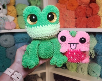 Op de afbeelding: Twee gehaakte knuffels, een groene kikker en een roze aardbei-vormig wezen met zijn tong naar buiten. De kikker heeft grote gouden ogen en een groen-wit lichaam. Het aardbeiwezen heeft roze-witte strepen en een rood aardbeidesontwerp. Beide zitten op een plank met kleurrijke garenbollen.