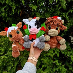 May include: Three handmade crochet plush toys: a gingerbread cow, a black and white cow, and a brown cow. Each plush has a festive holiday theme with red and green accents. The toys are held in front of a green leafy background.