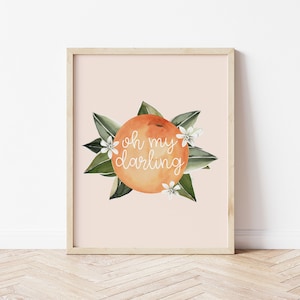 May include: A framed watercolor print featuring an orange with the words "oh my darling" in white script. The orange is surrounded by green leaves and white flowers, set against a pale peach background.