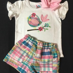 Pu&ograve; includere: Una maglietta bianca con maniche a balze e un'applicazione a forma di uccello, abbinata a pantaloncini a quadri. L'uccello &egrave; posato su un ramo con fiori. Un grande fiocco rosa &egrave; posto sopra la maglietta. I pantaloncini presentano un motivo a quadri patchwork.