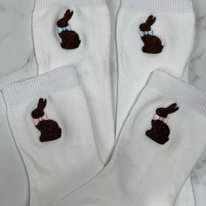 K&ouml;nnte beinhalten: Vier wei&szlig;e Socken mit gestickten braunen Schokoladenhasen-Designs. Jeder Hase hat eine hellblaue Schleife.