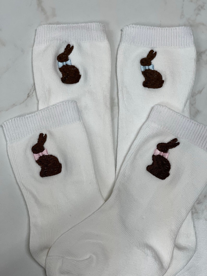 K&ouml;nnte beinhalten: Vier wei&szlig;e Socken mit gestickten braunen Schokoladenhasen-Designs. Jeder Hase hat eine hellblaue Schleife.