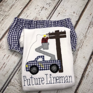 May include: White onesie with a blue and white checkered truck, a lineman in a bucket truck, and the text "Future Lineman". The onesie is paired with blue and white checkered bloomers.