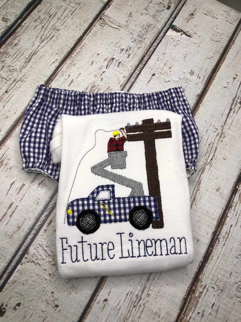 May include: White onesie with a blue and white checkered truck, a lineman in a bucket truck, and the text "Future Lineman". The onesie is paired with blue and white checkered bloomers.