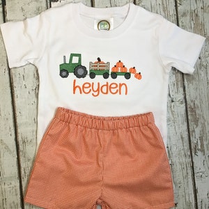 May include: White short-sleeved t-shirt with an embroidered green tractor pulling a wagon full of orange pumpkins. The shirt has the name "heyden" embroidered below the tractor. The shirt is paired with orange and white gingham shorts.
