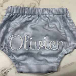 Op de afbeelding: Babybroekje in blauw en wit ruitjespatroon, met een elastische tailleband. De naam "Olivier" is in witte letters geborduurd. Een schattig en comfortabel babykledingstuk.