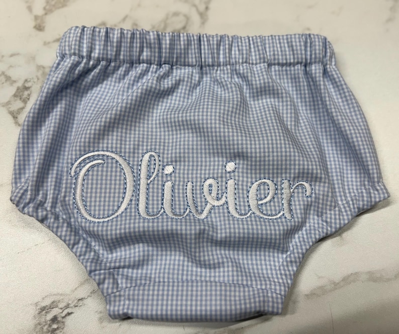 Op de afbeelding: Babybroekje in blauw en wit ruitjespatroon, met een elastische tailleband. De naam "Olivier" is in witte letters geborduurd. Een schattig en comfortabel babykledingstuk.