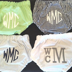 Op de afbeelding: Vier babybloomers in verschillende patronen en kleuren. De bloomers zijn onder andere groen ruitjes, marineblauw en wit ruitjes, blauw-witte strepen en wit. Elke broek heeft een monogram in een contrasterende kleur.