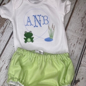 May include: White baby onesie with blue embroidered initials, a green frog, and a pond scene. Paired with green and white gingham bloomers. The onesie has the word "child" on the tag.