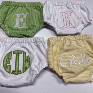 Op de afbeelding: Vier babybloomers in groene gingham, witte en geel gestreepte stoffen. Elke bloomer heeft geborduurde letters: een "E", een monogram en de naam "Estelle". De bloomers hebben elastische taillebanden.
