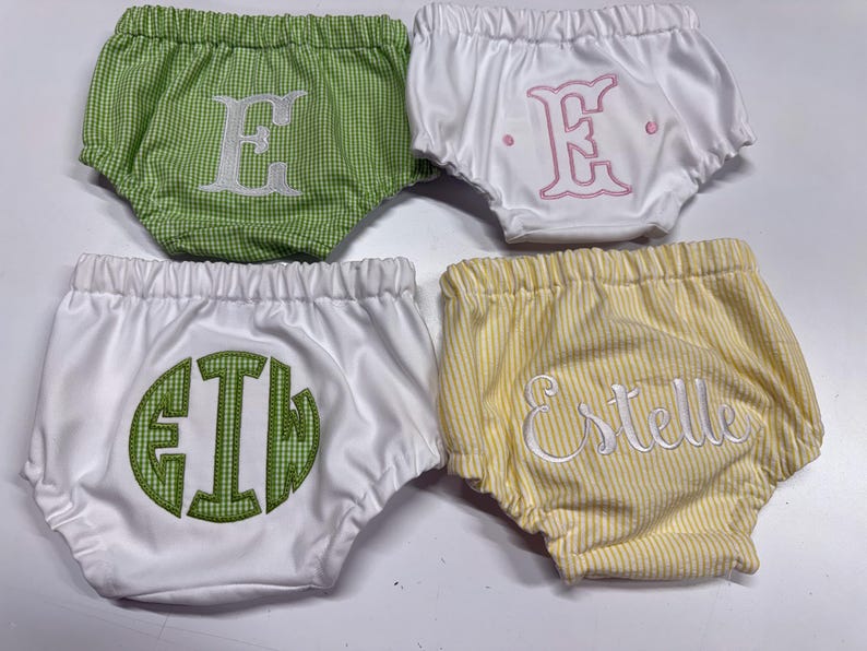 Op de afbeelding: Vier babybloomers in groene gingham, witte en geel gestreepte stoffen. Elke bloomer heeft geborduurde letters: een "E", een monogram en de naam "Estelle". De bloomers hebben elastische taillebanden.