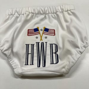 Op de afbeelding: Witte babybloomers met een elastische tailleband. De bloomers hebben een geborduurd ontwerp van twee gekruiste Amerikaanse vlaggen, sterren en de letters "HWB" in marineblauw.