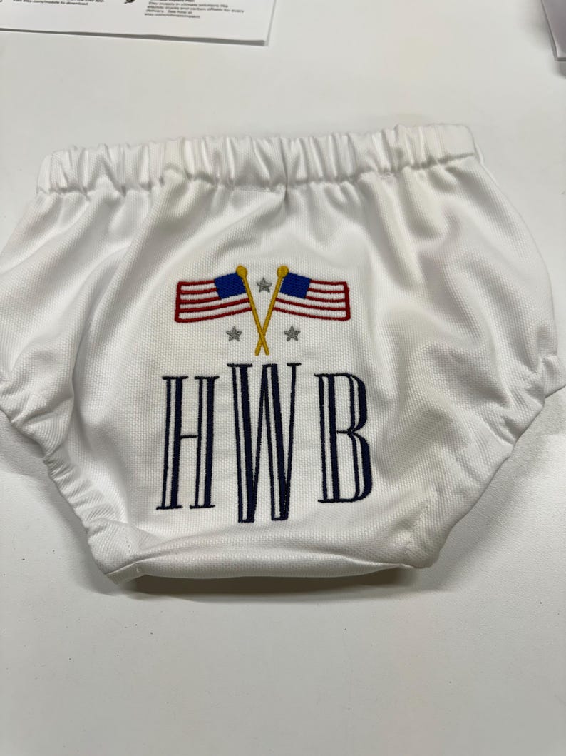 Op de afbeelding: Witte babybloomers met een elastische tailleband. De bloomers hebben een geborduurd ontwerp van twee gekruiste Amerikaanse vlaggen, sterren en de letters "HWB" in marineblauw.