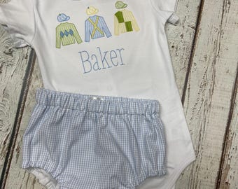 Puede incluir: Conjunto de body blanco y cubrepañales de cuadros azules para bebé. El body presenta sedas de jockey bordadas y el nombre "Baker" en hilo azul. El conjunto es para un bebé.