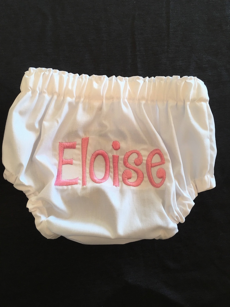 Op de afbeelding: Witte babybloomers met elastische taille en beenopeningen. De naam "Eloise" is roze op de voorkant geborduurd. De bloomer is gemaakt van een zachte, lichte stof.