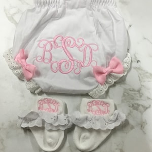 May include: White baby bloomers with pink embroidered initials "BSD" and white lace trim. The bloomers are paired with white socks with pink embroidered initials "BSD" and white lace trim.