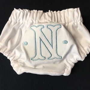 Op de afbeelding: Witte babybloomers met een elastische tailleband en beenopeningen. De bloomers hebben een grote, geborduurde letter "N" in lichtblauw op de achterkant, met twee kleine geborduurde stippen aan weerszijden.