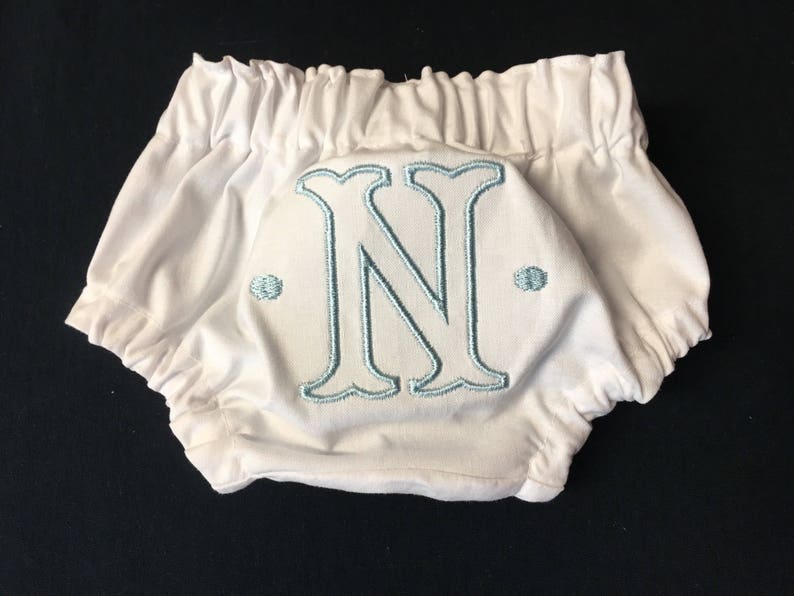 Op de afbeelding: Witte babybloomers met een elastische tailleband en beenopeningen. De bloomers hebben een grote, geborduurde letter "N" in lichtblauw op de achterkant, met twee kleine geborduurde stippen aan weerszijden.