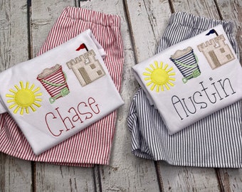 May include: Two white t-shirts with red and blue striped pants. The shirts have embroidered images of a sun, a bucket and a sandcastle. The shirts are personalized with the names "Chase" and "Austin".