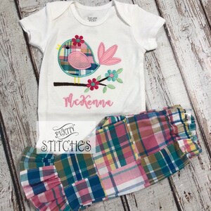 Pu&ograve; includere: Un body bianco per neonati con un'applicazione a forma di uccello, con dettagli rosa, blu e verdi, e il nome "McKenna" ricamato in rosa. Abbinato a pantaloni patchwork a quadri con polsini a balze. Il marchio "Purity Stitches" &egrave; visibile.