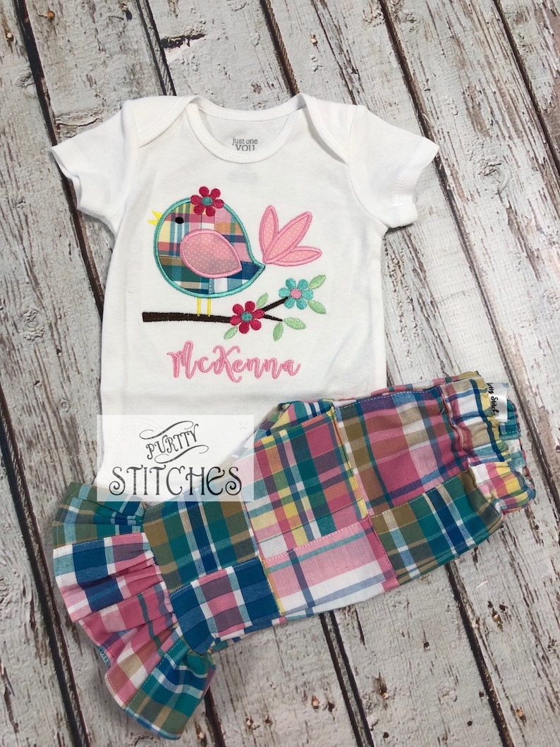 Pu&ograve; includere: Un body bianco per neonati con un'applicazione a forma di uccello, con dettagli rosa, blu e verdi, e il nome "McKenna" ricamato in rosa. Abbinato a pantaloni patchwork a quadri con polsini a balze. Il marchio "Purity Stitches" &egrave; visibile.