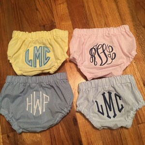 Op de afbeelding: Vier babybloomers in verschillende kleuren en patronen. E&eacute;n is geel met blauwe borduursels, een andere roze met blauwe borduursels, een blauw geruit paar met witte borduursels en een blauw-wit gestreept paar met blauwe borduursels. Elk paar heeft monograminitialen.